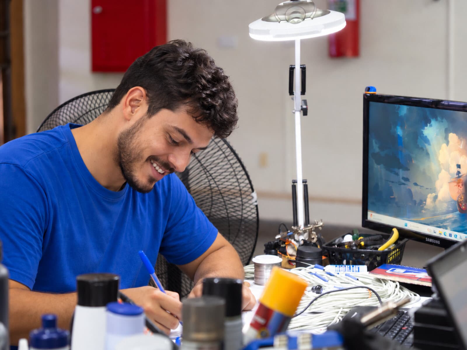 Técnico na bancada de montagem da PETE, com luminária de lupa e monitor