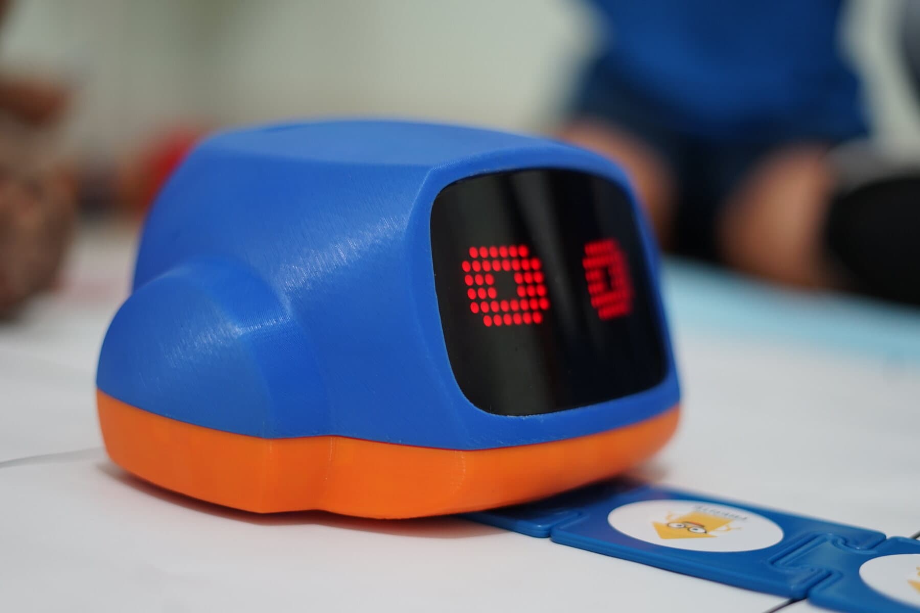 Robô PETE ROB em três quartos, carcaça azul com base laranja e visor de matriz de LED, sobre o tapete de atividades
